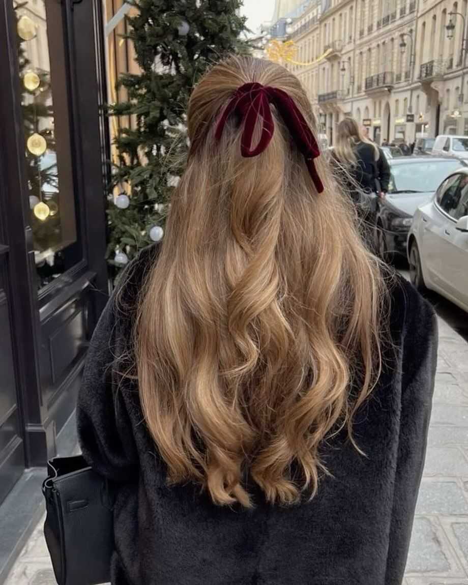 Woman with wavy hair tied with a velvet bow, walking on a city street decorated for the holidays.